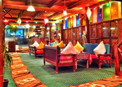 A quaint restaurant full of colored glass windows and benches covered in colorful pillows.